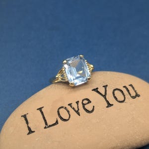 May include: A gold ring with a large, rectangular, light blue gemstone. The ring is set on a tan stone engraved with the words "I Love You". The background is a solid, dark blue.