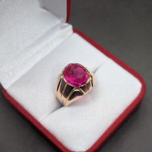May include: A gold ring with a large, oval-shaped, pink gemstone set in the center. The ring is in a red velvet ring box.