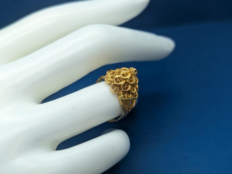 May include: A gold filigree ring with a detailed design. The ring is worn on a model's finger.