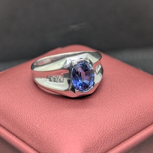 May include: A silver ring with a large, oval, blue gemstone at the center. The ring has a rectangular design with small accent stones on each side of the main stone. The ring is displayed on a red surface.