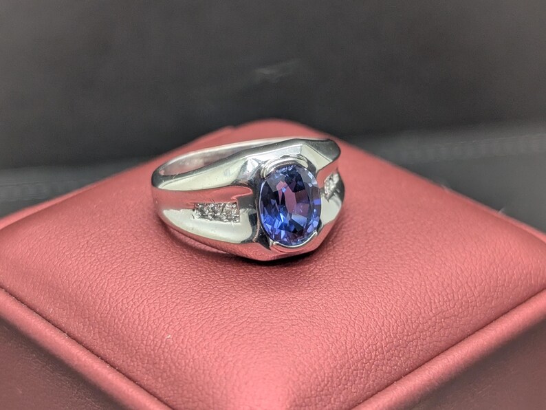 May include: A silver ring with a large, oval, blue gemstone at the center. The ring has a rectangular design with small accent stones on each side of the main stone. The ring is displayed on a red surface.