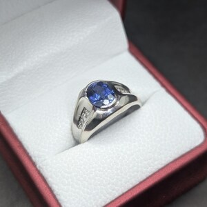 May include: A silver ring with a large oval blue gemstone and small accent diamonds. The ring is set in a white box with a red exterior, against a dark background. The ring's design is classic and elegant.