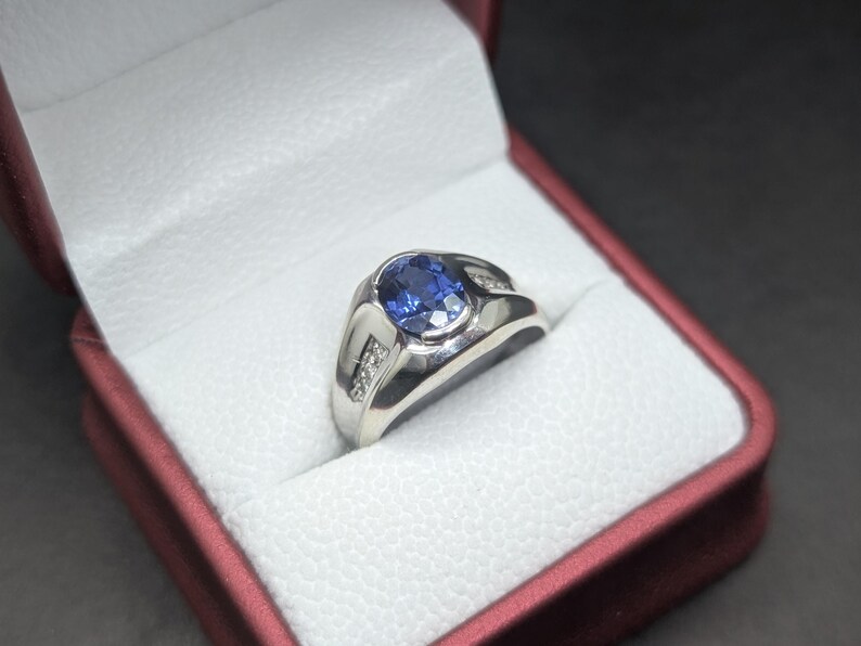 May include: A silver ring with a large oval blue gemstone and small accent diamonds. The ring is set in a white box with a red exterior, against a dark background. The ring's design is classic and elegant.