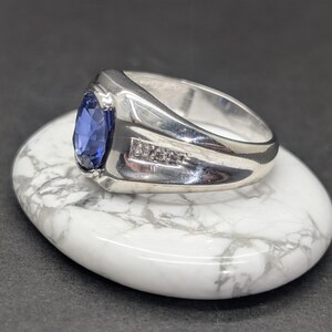 May include: A silver ring with a large, oval, blue gemstone and small accent stones. The ring is set on a white and gray marbled stone, with a dark background.