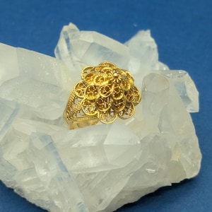 May include: A gold filigree ring with a flower-shaped design. The ring is set on a bed of white crystals.