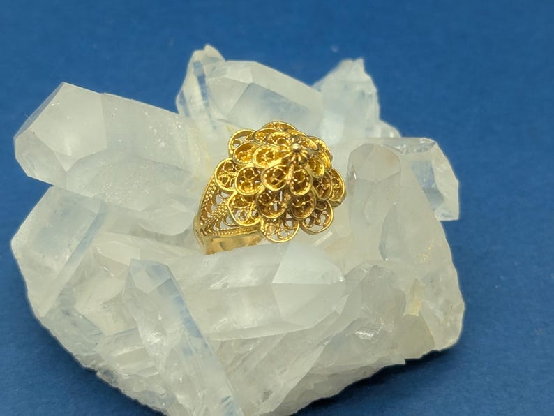 May include: A gold filigree ring with a flower-shaped design. The ring is set on a bed of white crystals.