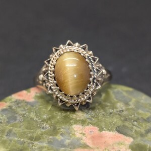 May include: A vintage ring with a large, oval, golden-brown gemstone. The stone is set in a silver-toned metal with a decorative, sunburst-style setting. The ring is displayed on a green and pink stone surface.
