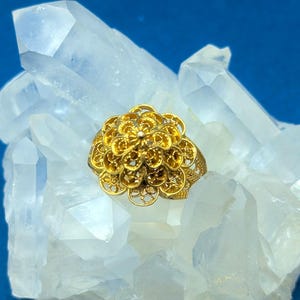 May include: A gold filigree ring with a flower design. The ring is sitting on a bed of white quartz crystals.