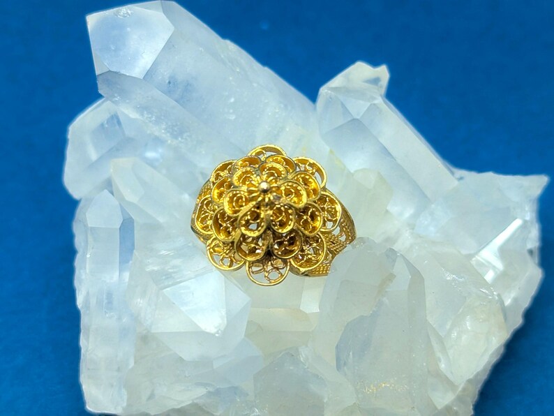 May include: A gold filigree ring with a flower design. The ring is sitting on a bed of white quartz crystals.