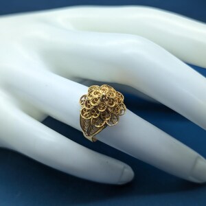 May include: A gold filigree ring with a delicate floral design. The ring is on a white mannequin hand.