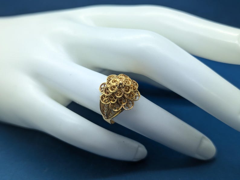 May include: A gold filigree ring with a delicate floral design. The ring is on a white mannequin hand.