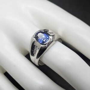 May include: A silver ring with a large oval blue gemstone at the center. The ring has a modern design with small accent stones on each side of the main stone. The ring is displayed on a white surface.