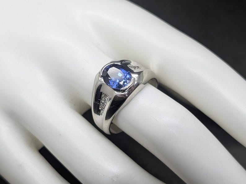 May include: A silver ring with a large oval blue gemstone at the center. The ring has a modern design with small accent stones on each side of the main stone. The ring is displayed on a white surface.