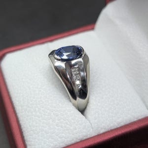 May include: A silver ring with a large oval blue gemstone and three smaller accent stones. The ring is set in a polished silver band and is displayed in a red and white ring box. The ring is a classic design.