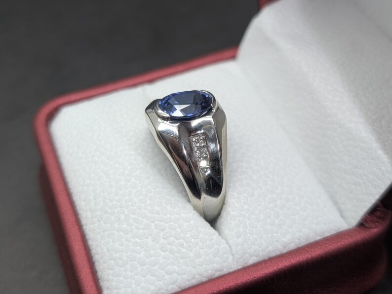 May include: A silver ring with a large oval blue gemstone and three smaller accent stones. The ring is set in a polished silver band and is displayed in a red and white ring box. The ring is a classic design.