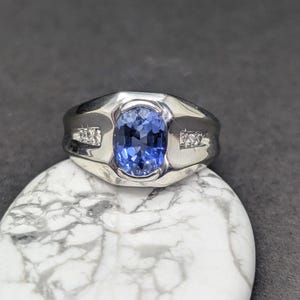 May include: A silver ring featuring a large, oval-cut blue gemstone as the centerpiece. The ring has a wide band and is accented with small, clear stones on each side of the main stone. The ring is displayed on a white and gray marbled surface.