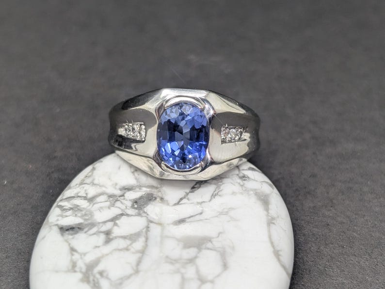 May include: A silver ring featuring a large, oval-cut blue gemstone as the centerpiece. The ring has a wide band and is accented with small, clear stones on each side of the main stone. The ring is displayed on a white and gray marbled surface.