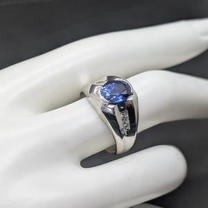 May include: A silver ring with a large oval blue gemstone and two smaller clear stones on each side. The ring is displayed on a white hand against a dark background. The ring's design is classic and elegant.