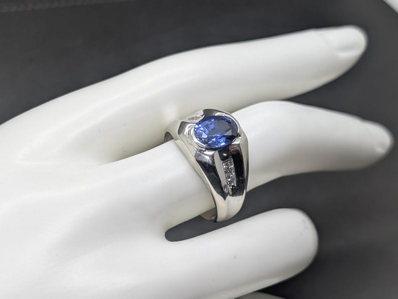 May include: A silver ring with a large oval blue gemstone and two smaller clear stones on each side. The ring is displayed on a white hand against a dark background. The ring's design is classic and elegant.