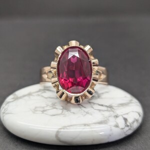 May include: A gold-colored ring with a large, oval, ruby-red gemstone. The ring's setting has a decorative, scalloped design around the gemstone. The ring is displayed on a white, marble-patterned surface against a dark background.