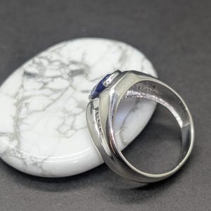 May include: A silver ring with a dark blue gemstone and small accent stones. The ring is set on a white, oval-shaped stone with gray marbling. The background is a dark gray surface.