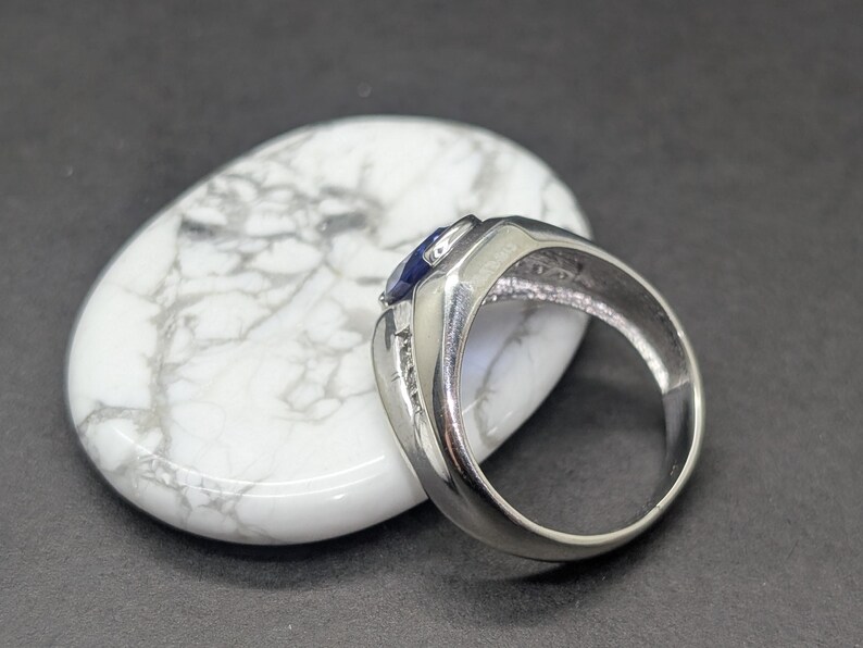May include: A silver ring with a dark blue gemstone and small accent stones. The ring is set on a white, oval-shaped stone with gray marbling. The background is a dark gray surface.