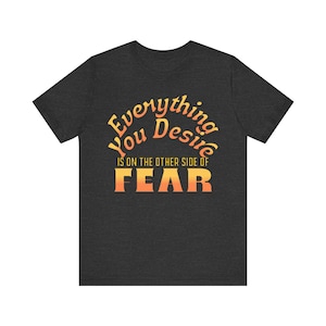 May include: A dark charcoal grey short-sleeve t-shirt featuring the text Everything You Desire IS ON THE OTHER SIDE OF FEAR in a gradient orange and yellow colour scheme with a horizontal line pattern across the letters of the word FEAR.