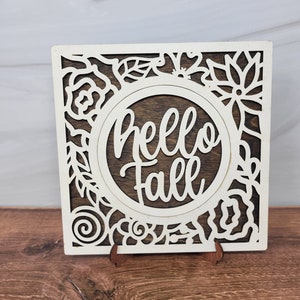 May include: White wooden sign with a brown wood background and a cutout design of flowers and leaves. The sign says "Hello Fall" in a white script font.