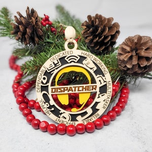 Puede incluir: Un adorno navideño de madera amarillo y negro con la palabra "DISPATCHER" en texto marrón. El adorno está rodeado por un círculo de palabras que describen a un despachador, incluyendo "fiable", "dedicado", "trabajador", "multitarea", "fiable", "servicial" y "eficiente".