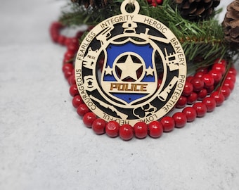 Könnte beinhalten: Ein hölzerner Weihnachtsbaumschmuck in Form eines Polizeibadgens mit dem Wort "POLICE" in der Mitte. Der Anhänger ist von einem Kreis aus Wörtern umgeben, die positive Eigenschaften beschreiben, wie "Integrität", "Heroisch", "Tapfer", "Schützend", "Mutig", "Furchtlos", "Loyal", "Hilfsbereit".
