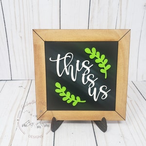 Könnte beinhalten: Ein kleines, mit Holz gerahmtes Schild mit einer schwarzen Tafel, auf der der Text "This is us" in weißen Buchstaben mit grünen Blatt-Akzenten steht.