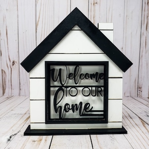 May include: White wooden house shaped sign with black trim and a black cutout of the words "Welcome to our home" in the centre.