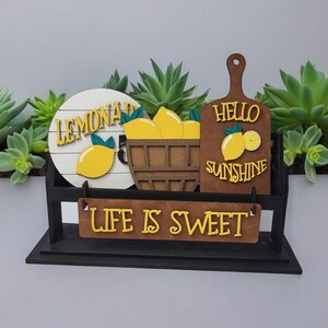 May include: Wooden sign with the words "Lemonade", "Hello Sunshine", and "Life is Sweet" in yellow letters. The sign is decorated with lemons and a basket of lemons.