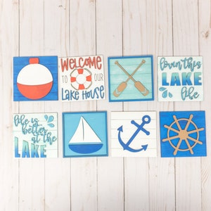 Puede incluir: Conjunto de ocho letreros de madera con temática náutica. Los letreros presentan varios diseños, incluyendo un salvavidas, un velero, un ancla, un timón de barco y el texto "Bienvenido a nuestra casa en el lago", "La vida es mejor en el lago" y "Amando esta vida en el lago".
