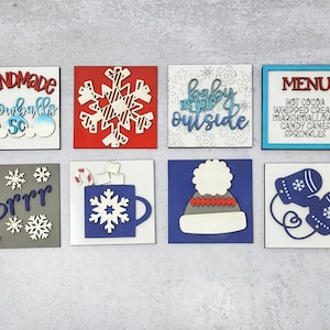 May include: Eight wooden signs with winter themes. The signs are painted in various colors, including red, blue, gray, and white. The signs feature snowflakes, a snowman, a mug of hot chocolate, mittens, and text that says "Handmade Snowballs 50c", "Brrr", "Baby It's Cold Outside", and "Menu: Hot Cocoa, Whipped Cream, Marshmallows, Candy Canes, Sprinkles".