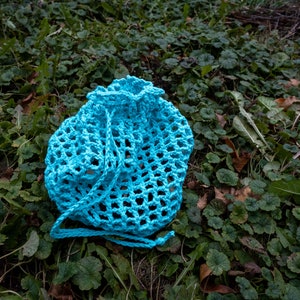May include: A light blue crocheted bag with a drawstring closure. The bag is lying on a bed of green leaves.