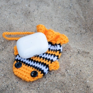 May include: A white wireless earbud case with a crocheted orange, black, and white fish-shaped cover. The case has a small loop for attaching to a keychain.