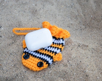 May include: A white wireless earbud case with a crocheted orange, black, and white fish-shaped cover. The case has a small loop for attaching to a keychain.