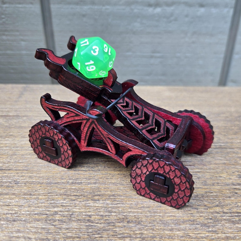 May include: A dark red wooden catapult with a dragon-inspired design and textured scale patterns on the wheels. The catapult features a black elastic band holding a bright translucent green twenty-sided die in the launching cradle.