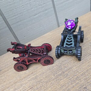 May include: Two miniature catapults with dragon-inspired frames. One is dark red with black accents, and the other is black with green highlights. The black catapult holds a purple twenty-sided die in its cradle, secured by a thin black elastic band.