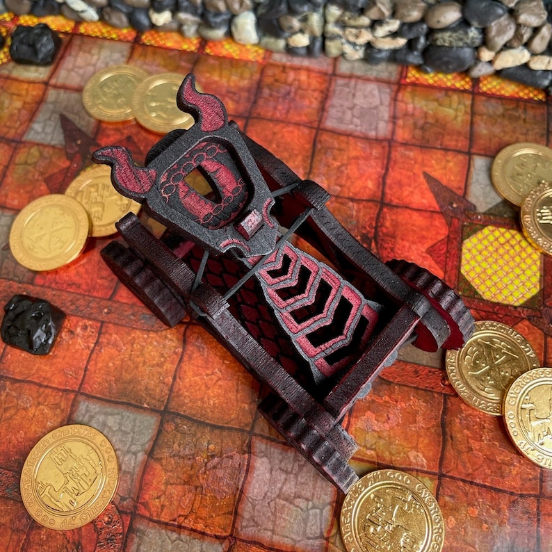 May include: A miniature wooden catapult featuring a dark brown frame with red accents and a horned design on the front. The device includes a central firing mechanism with a black elastic band and is surrounded by several gold-colored plastic coins.