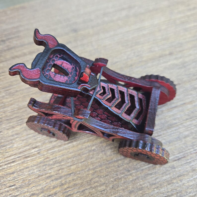 May include: A miniature wooden catapult model featuring a dark reddish-brown finish with black accents. The structure includes a central throwing arm, a tension-based elastic band mechanism, and four small gear-shaped wheels on the base.