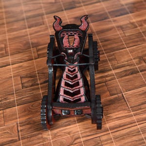 May include: A miniature black and red wooden catapult model featuring a horned top structure, a central tension band, and two side wheels. The frame is constructed with layered dark wood panels, showcasing a detailed geometric design.