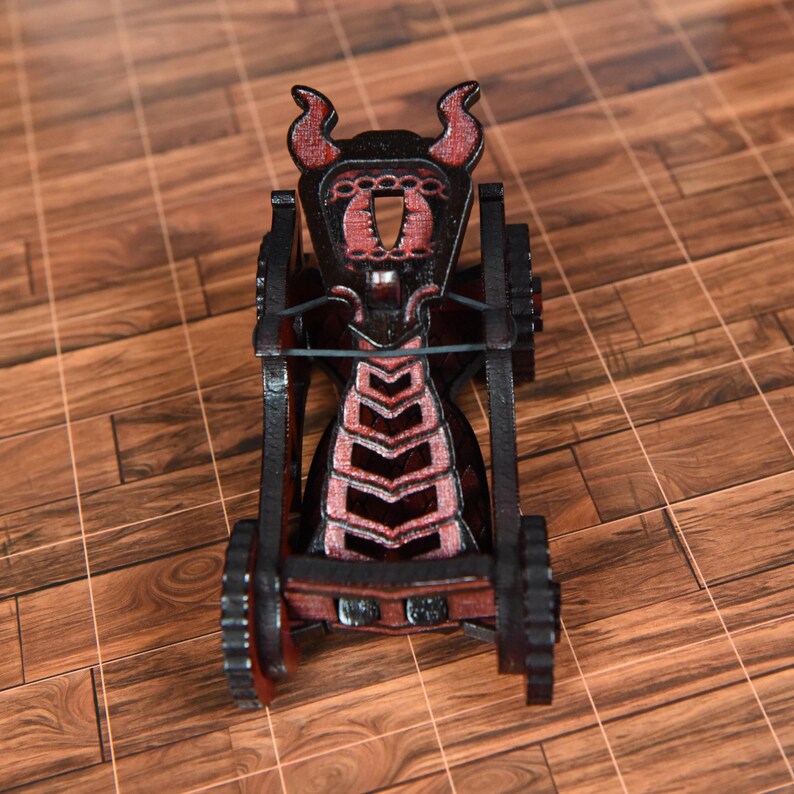 May include: A miniature black and red wooden catapult model featuring a horned top structure, a central tension band, and two side wheels. The frame is constructed with layered dark wood panels, showcasing a detailed geometric design.