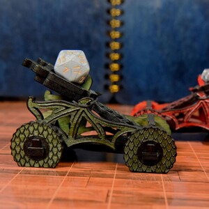 May include: A miniature green catapult with a textured scale pattern on the wheels and frame, holding a light gray twenty-sided die with gold numbers. The structure features intricate cutouts and a dark, weathered finish on the edges.