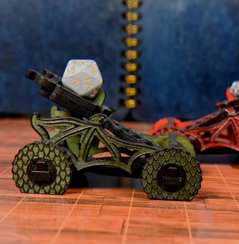 May include: A miniature green catapult with a textured scale pattern on the wheels and frame, holding a light gray twenty-sided die with gold numbers. The structure features intricate cutouts and a dark, weathered finish on the edges.