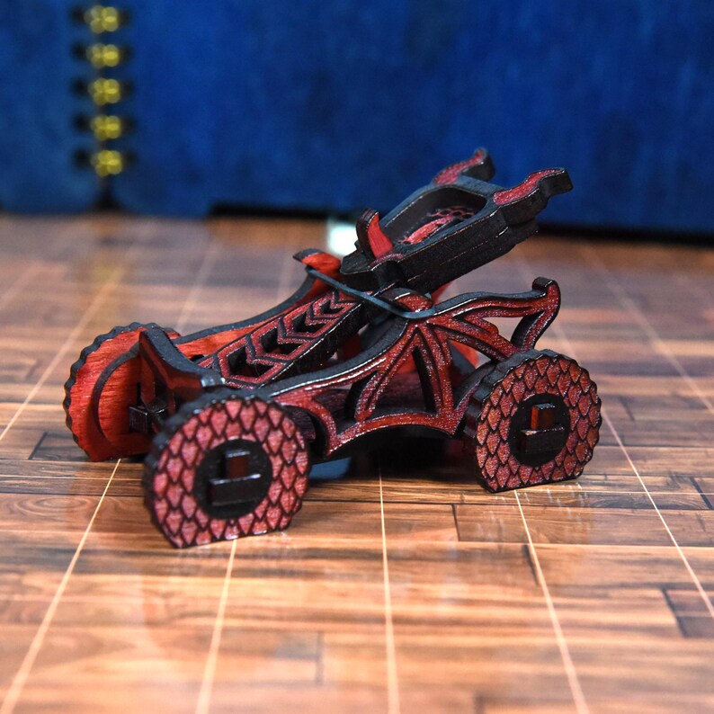 May include: A small, intricate wooden catapult model featuring a black and red color scheme. The structure includes dragon-wing side panels, wheels with a textured scale pattern, and a central firing mechanism held by a black elastic band.