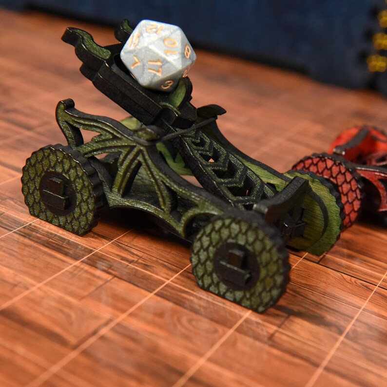 May include: A green and black wooden catapult toy featuring a dragon scale pattern on the wheels, holding a white twenty-sided die with gold numbers. A second red and black catapult is partially visible in the background to the right.