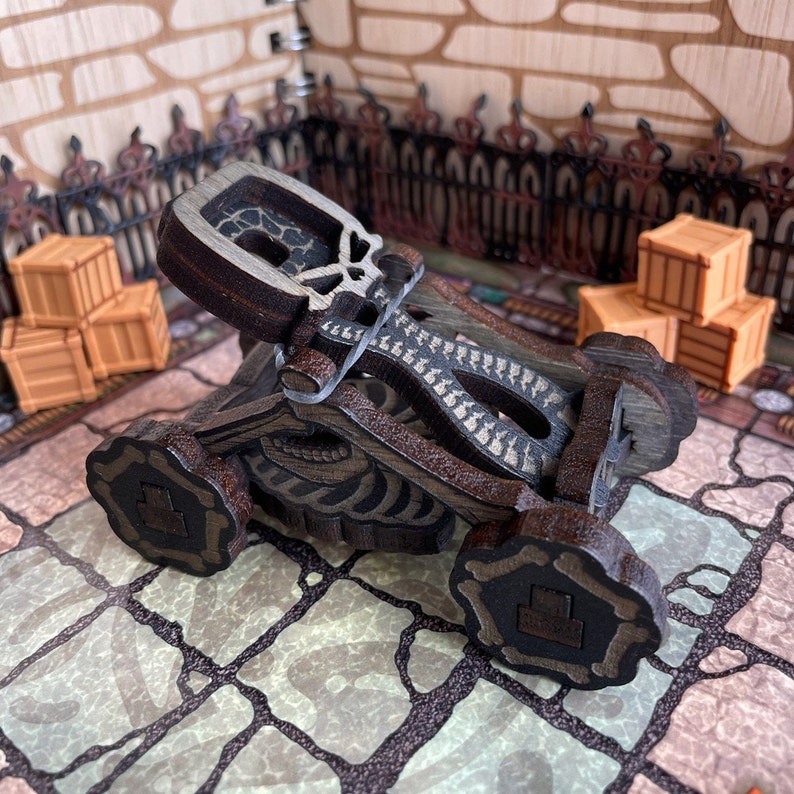 May include: A wooden skeletal catapult with a skull-shaped head, painted in brown and black with white accents. The catapult is on a stone-like surface.