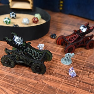 May include: Two miniature wooden catapults, one dark green and one dark red, each featuring a clear crystal projectile. A small gray figurine stands nearby on a brown grid surface, with a circular tray containing several multi-colored dice.